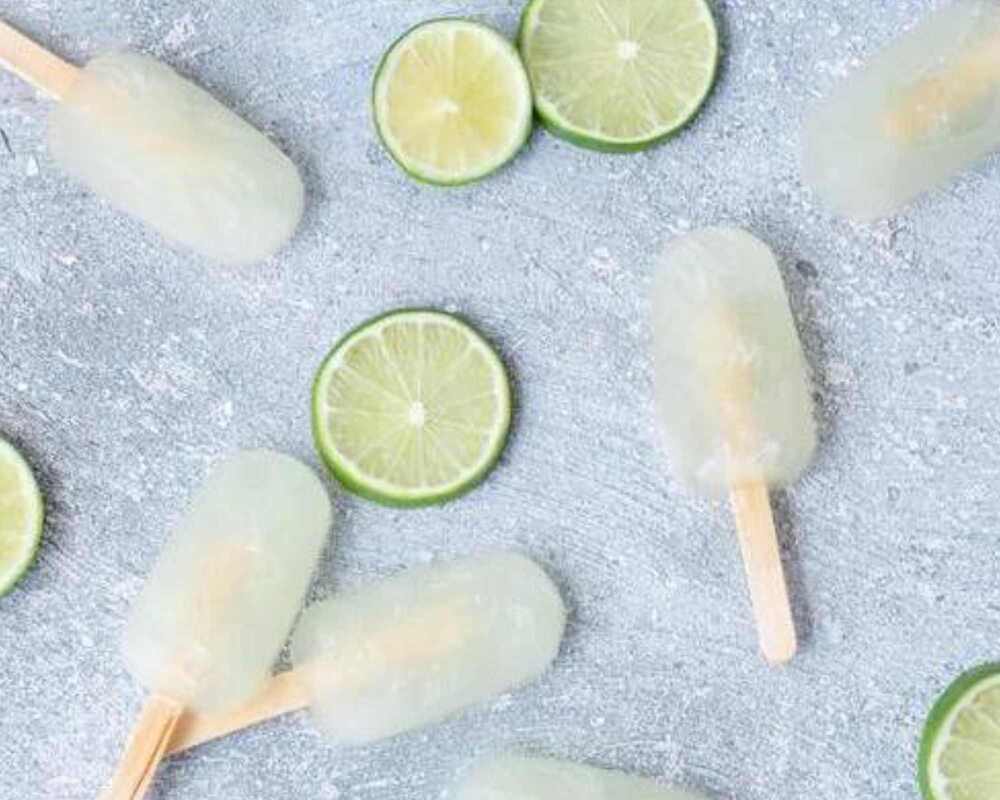 Margarita Popsicles on a grey table. They are surrounded by sliced limes.
