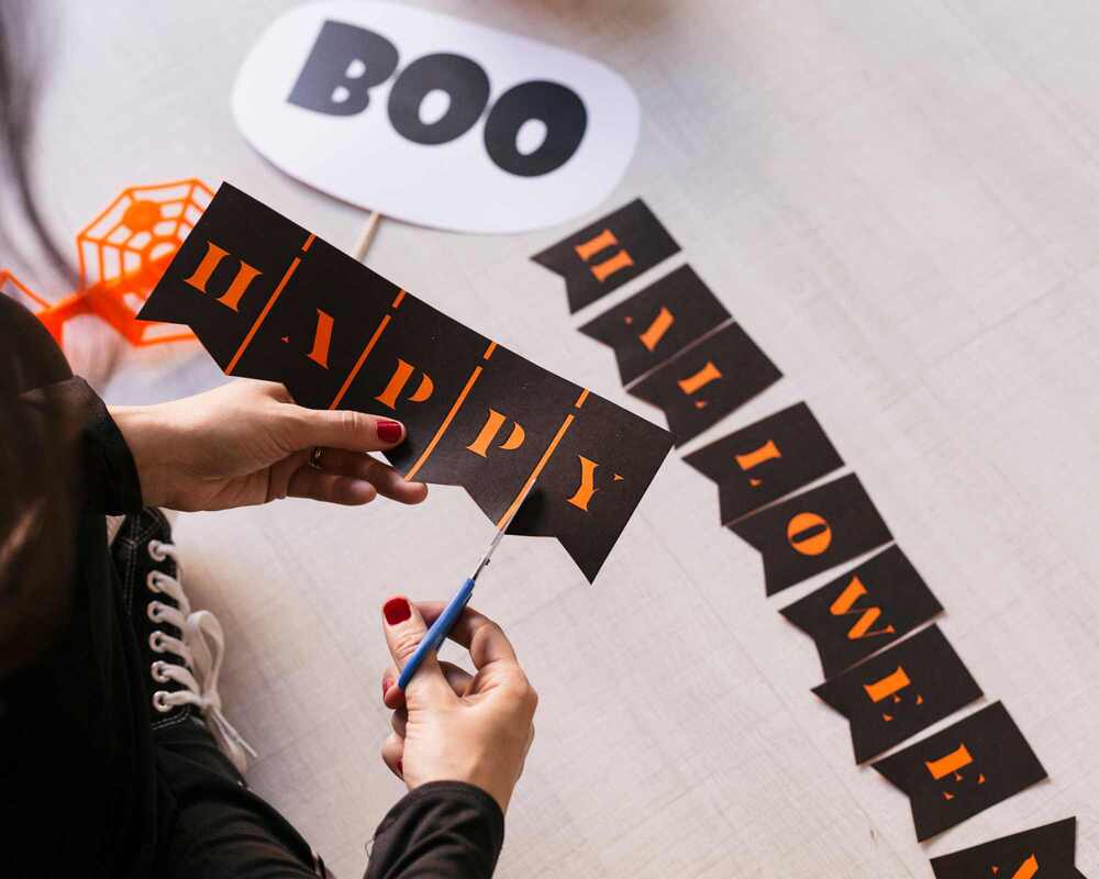 A person cutting out bunting that says 'Happy Halloween'. There is a sign that says 'boo' on the table.