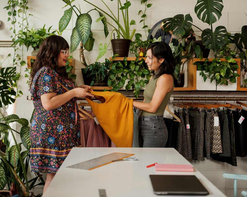A woman buying a yellow dress from another woman. They are standing in a shop and are surrounded by plants and sustainable clothing