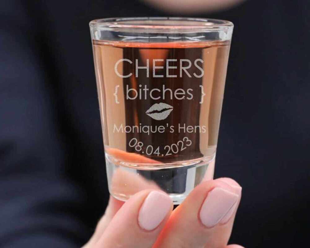 A hand holding a shot glass. Engraved on the shot glass is 'cheers bitches - Monique's hens 08.04.2023'