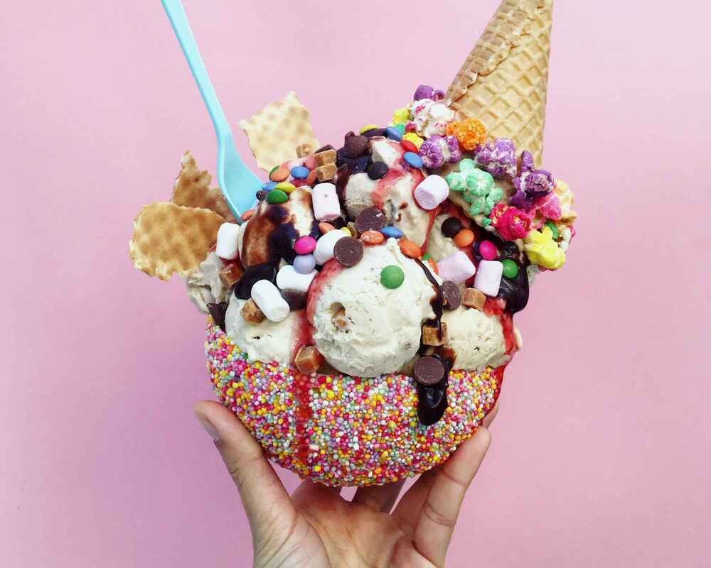 A person's hand holding a large ice-cream sundae topped with marshmallows, coloured popcorn, chocolates and lollies.