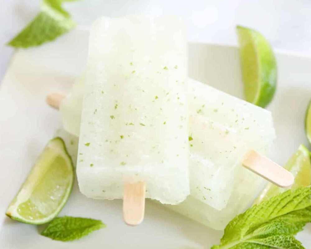 Two mojito popsicles on a plate. They are surrounded by cut up pieces of lime and mint leaves.