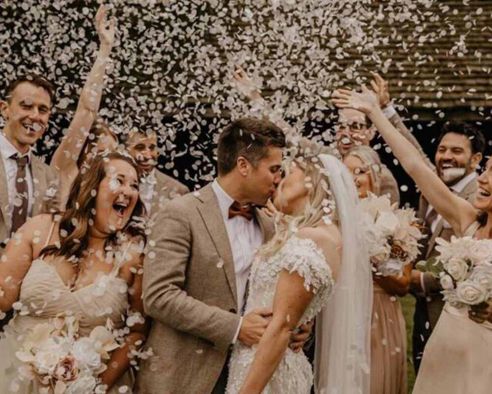 Newlyweds kissing, bridal party in the background throwing rice paper confetti in the air over the couple's heads