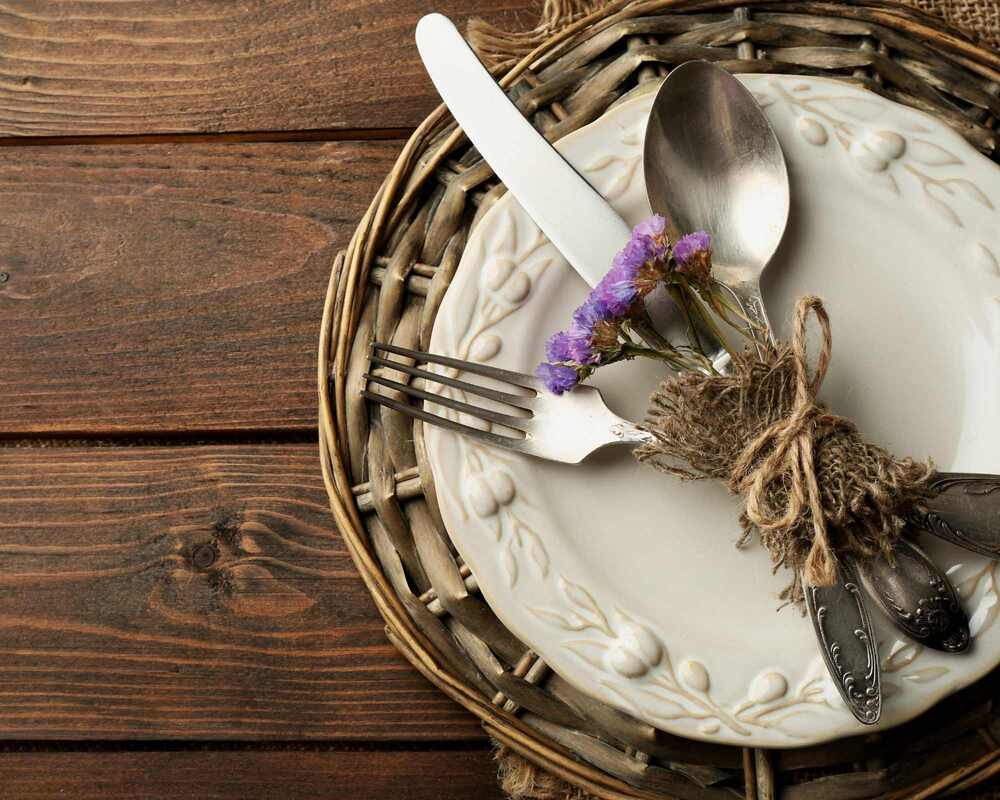 Wooden table with a white plate. There is cutlery tied together with twine and finished off with a small bunch of flowers
