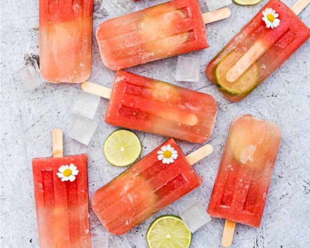 Frozen watermelon margarita popsicles. There are some on a steel tray, some on a steel plate and two pieces of watermelon on a white table