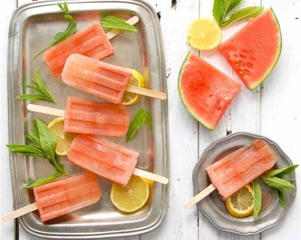 Watermelon mojitos placed on a stainless steel tray and plate. These are on a white table. Next to the mojitos is two slices of watermelon
